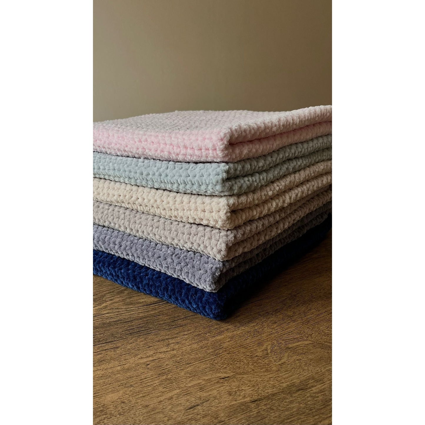 6 of the chunky blankets neatly sat on top of one another in the same way on a wooden table in a neutral space
